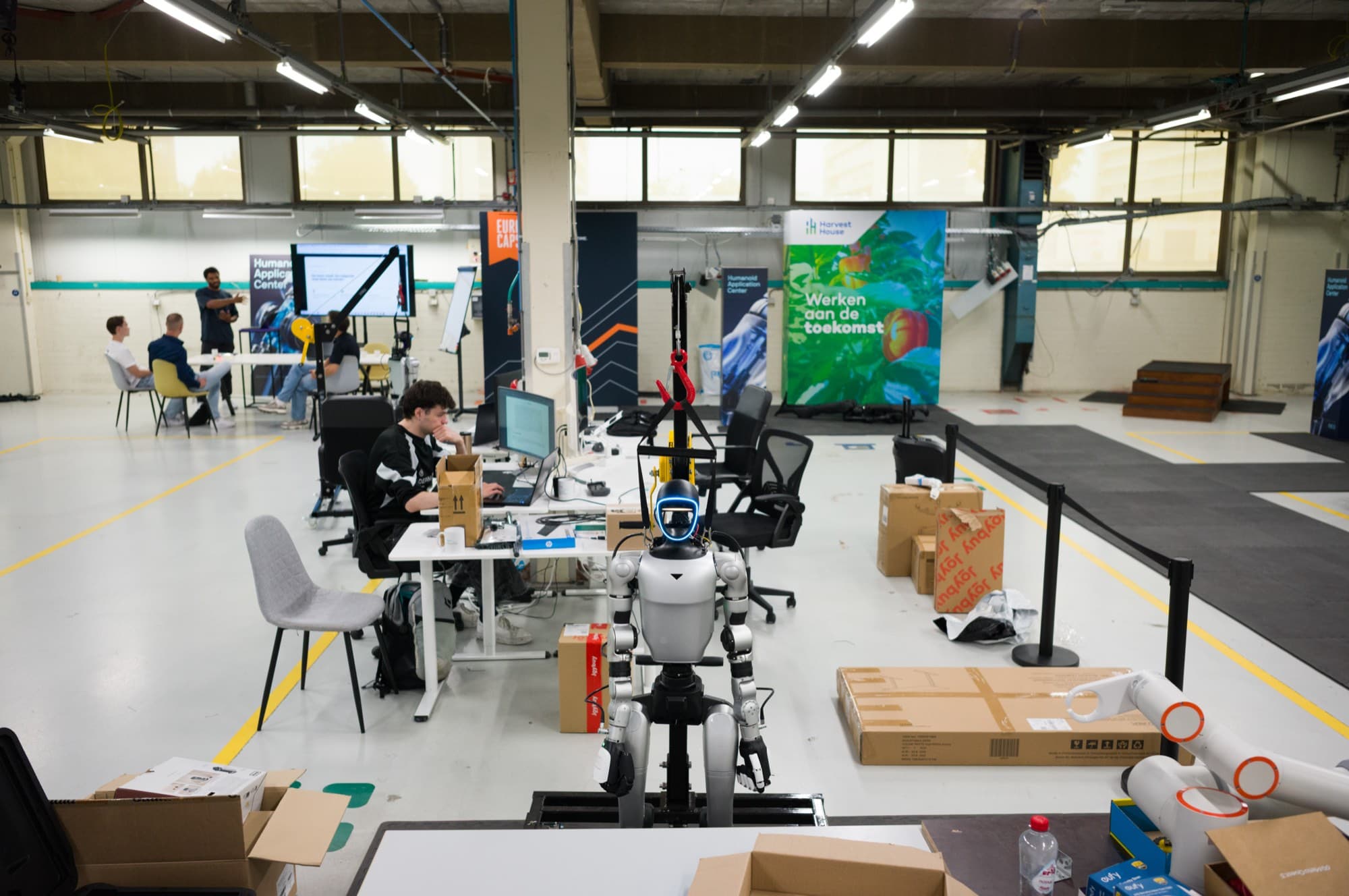The HAC shop floor with workstations, a humanoid in a hoist and a robot arm, while engineers are at work