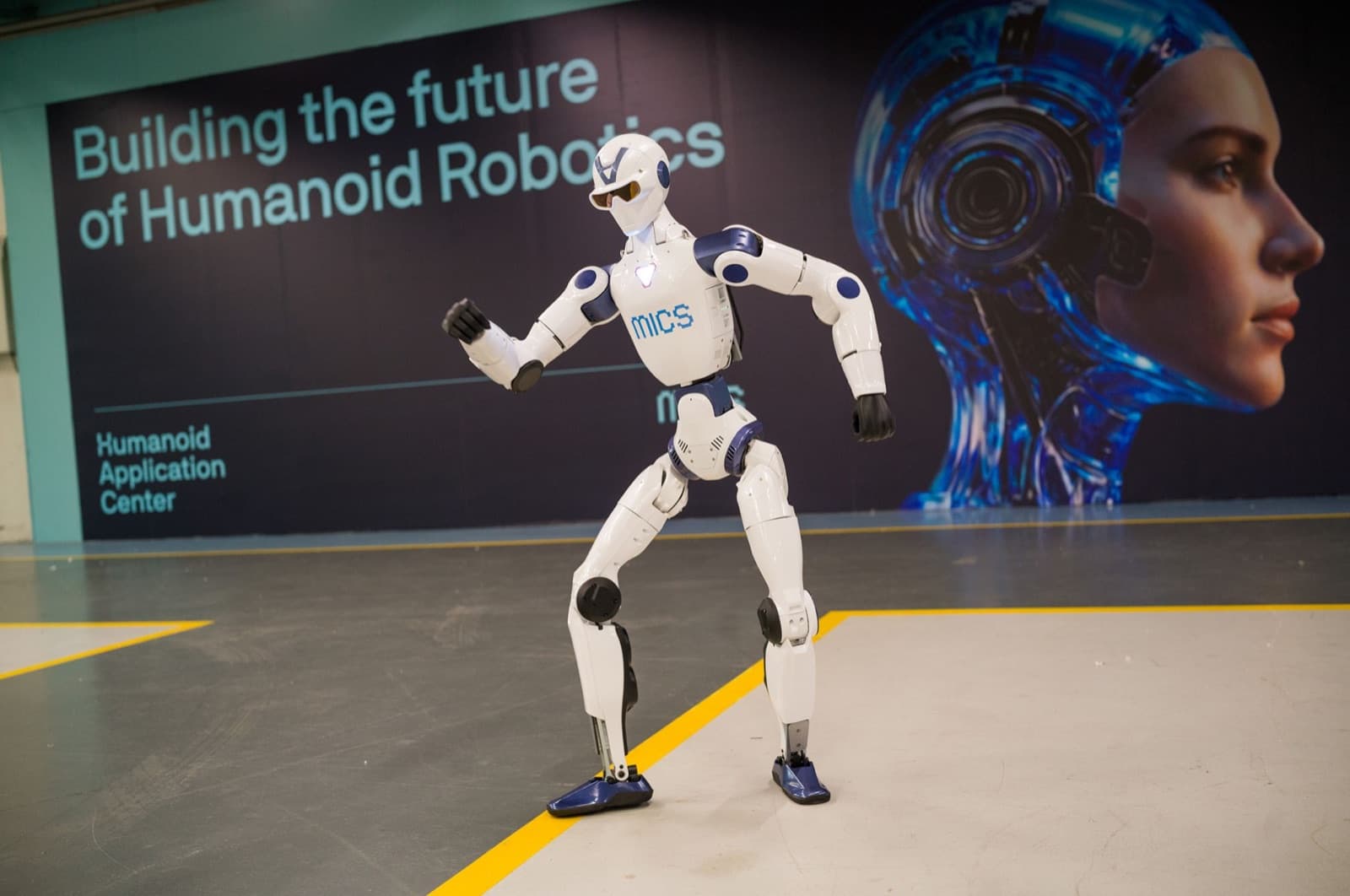 White MICS humanoid in a dynamic pose in front of the Building the future of Humanoid Robotics banner on the HAC floor