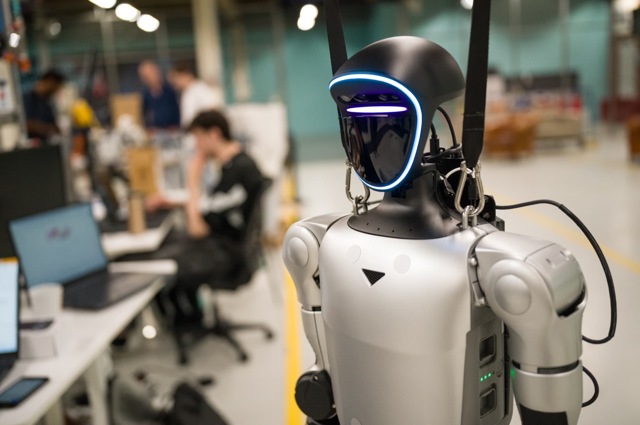 Humanoid robot met oplichtend vizier, op de achtergrond engineers aan het werk op de HAC-werkvloer