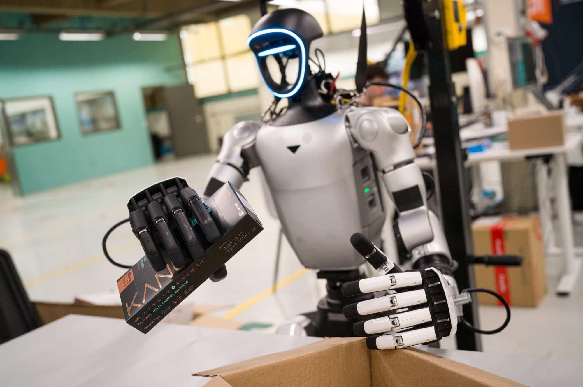 Humanoid picking up a small box and placing it in a shipping box on the HAC shop floor