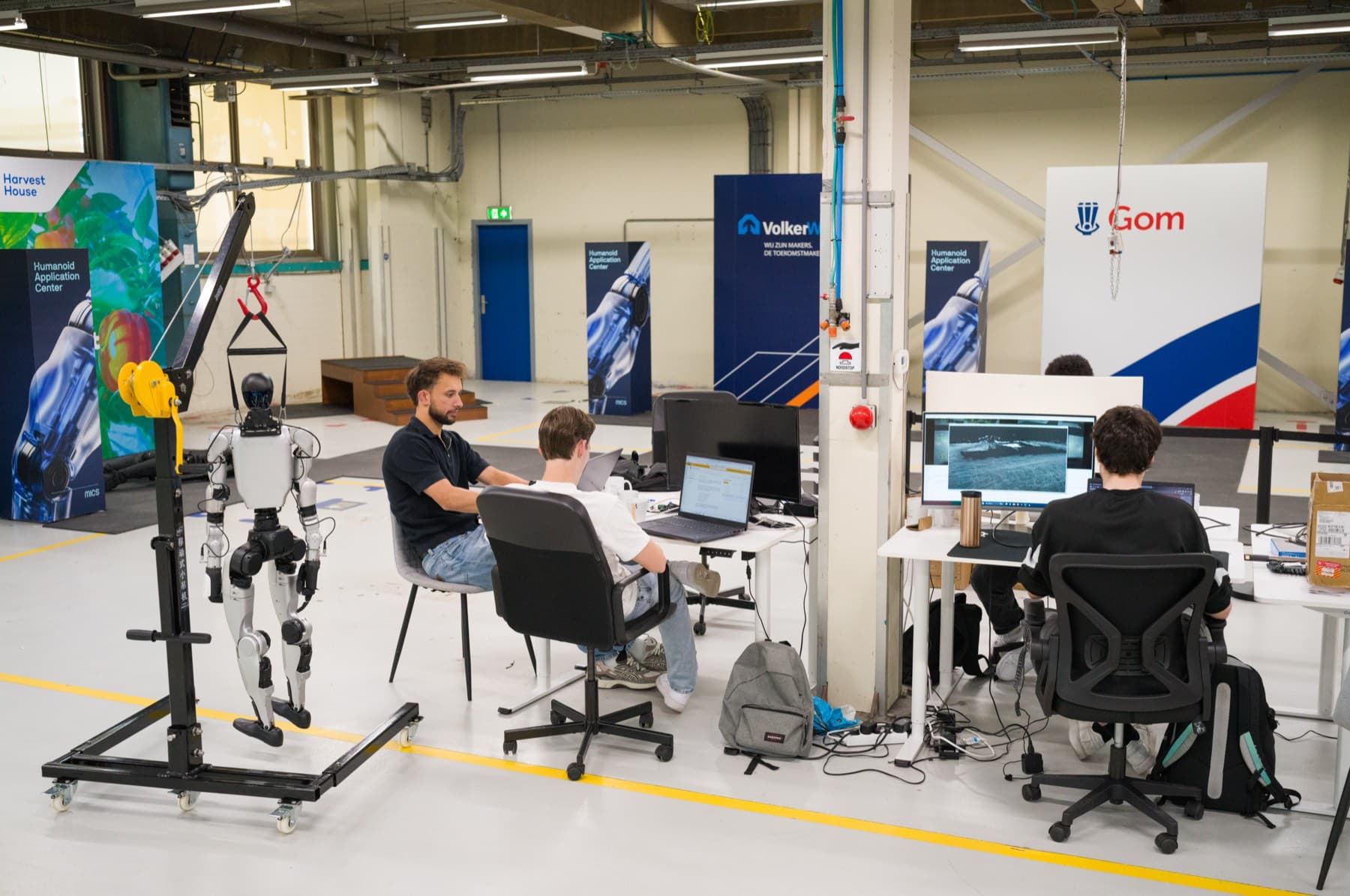 Engineers at their desks on the HAC shop floor, next to a humanoid in a hoist and the studio walls of Harvest House, VolkerWessels and Gom