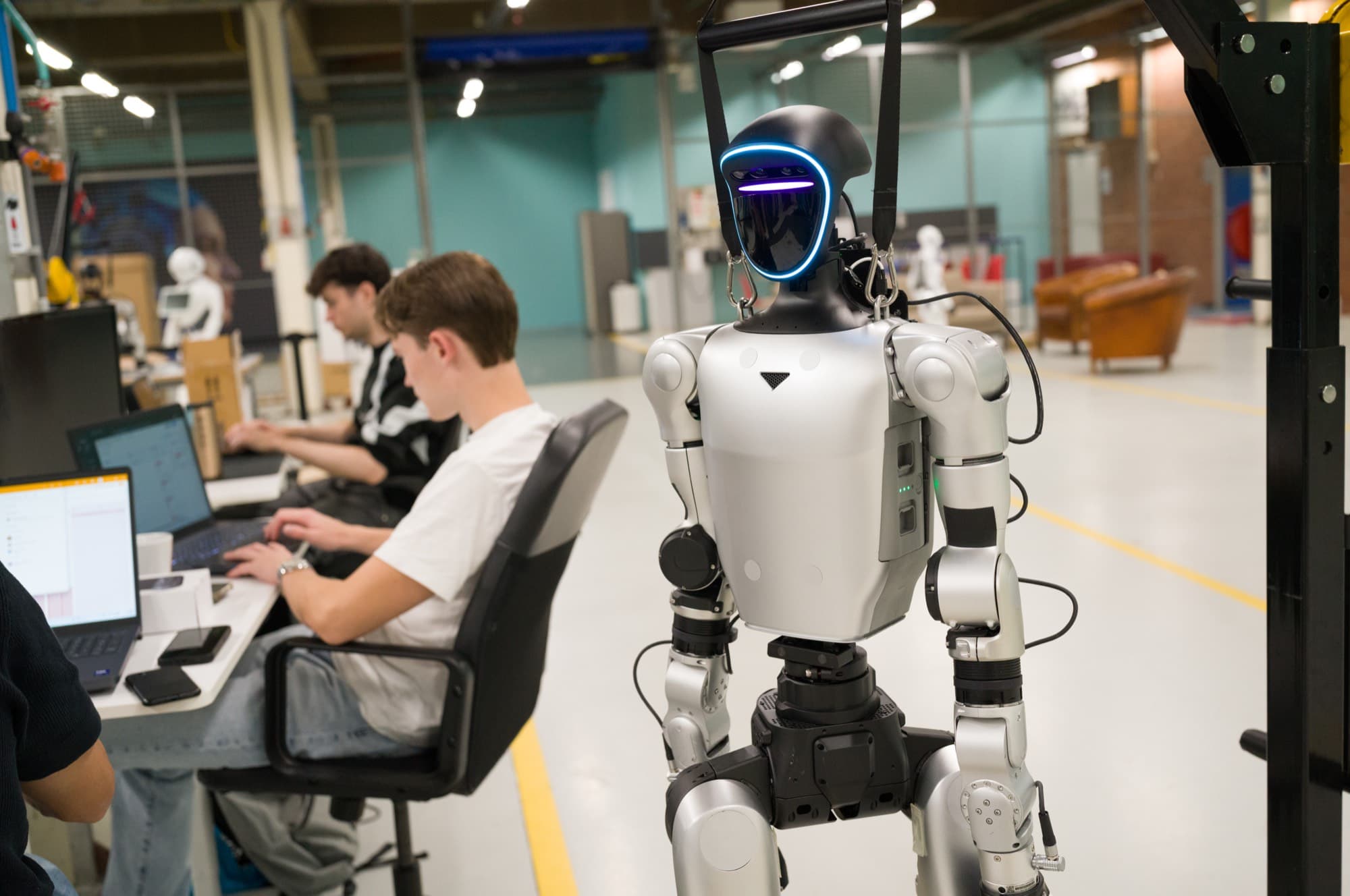 Twee engineers werken aan hun laptop op de HAC-werkvloer, naast een humanoid die in een takel hangt