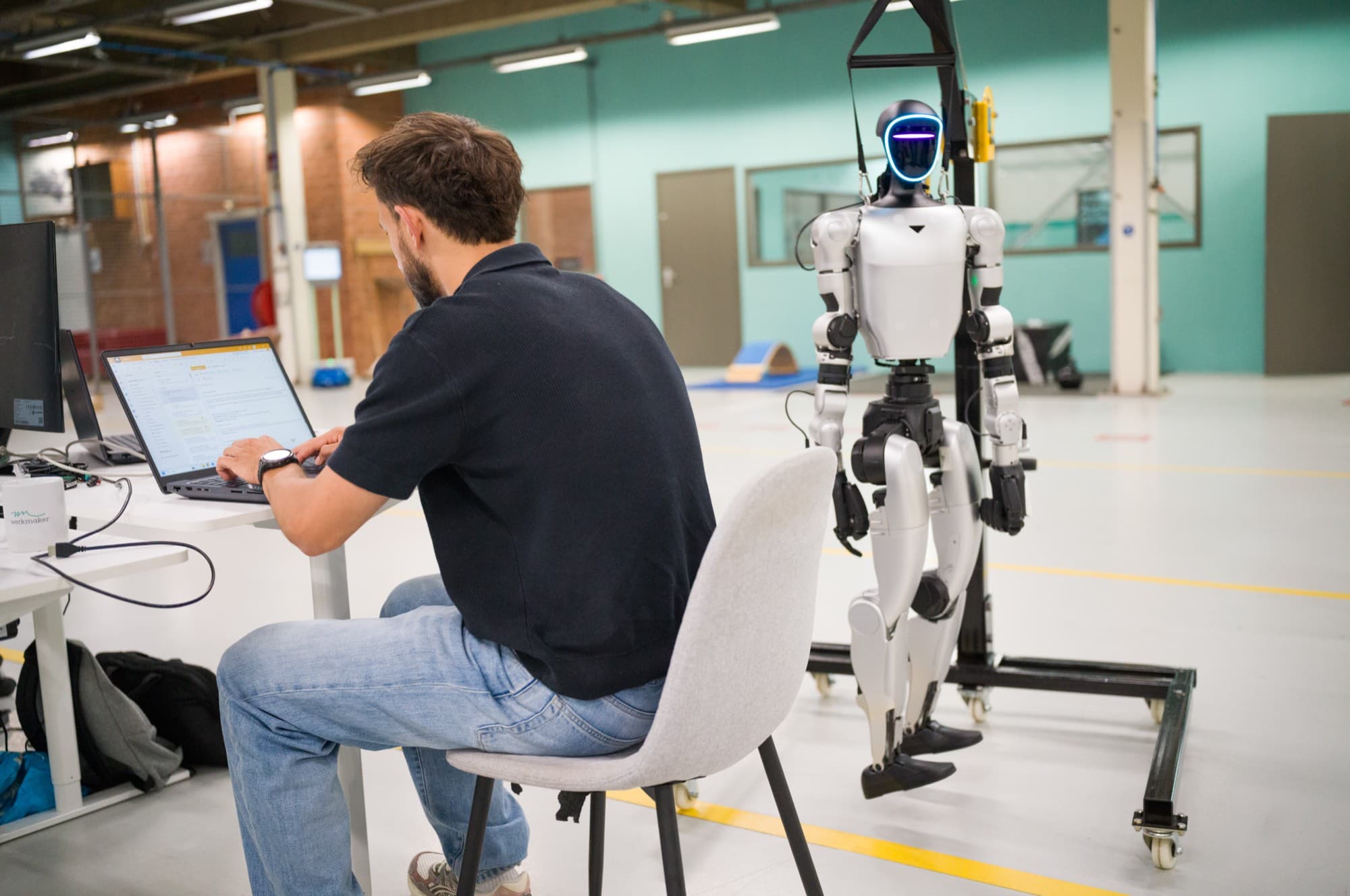 Engineer working on his laptop next to a humanoid hanging in a hoist on the HAC shop floor